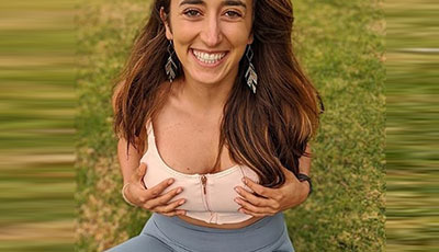 Sarafina Nance sitting on the grass smiling holding her breasts as she is wearing a compression sports bra